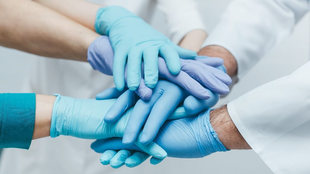 Team junger Ärzte, die in blauen Handschuhen in einem Innenraum ihre Hände übereinanderlegen. Medizinische Teamarbeit.