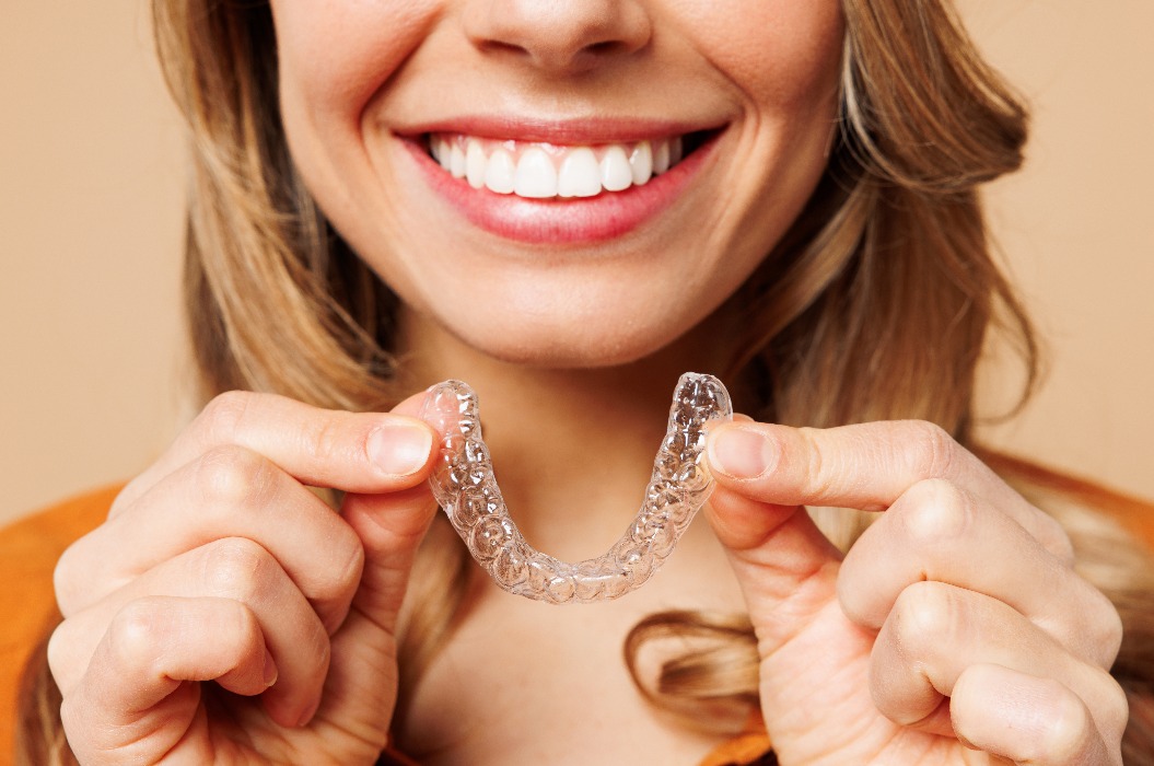 Nahaufnahme einer jungen Frau in orangefarbener Freizeitkleidung, die unsichtbare, transparente Aligner in der Hand hält; sie trägt eine Invisalign-Zahnspange und lächelt, isoliert vor einem schlichten, pastellfarbenen, hellbeigen Hintergrund. Lifestyle-Konzept.